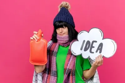 Young ill caucasian girl wearing winter hat and scarf wrapped in plaid holding idea bubble and hot water bag looking at hot water bag isolated on crimson background with copy space