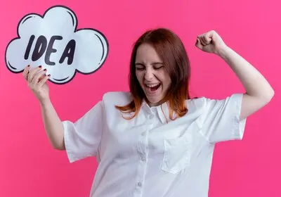 With closed eyes joyful young redhead girl holding idea bubble and raising fist isolated on pink wall