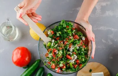 Top view woman making vegetable salad in glass bowl with tomatoes, cucumber, lemon on gray surface