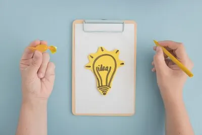 Hand holding yellow crayon and chocolate drawing light bulb on clipboard over the gray background
