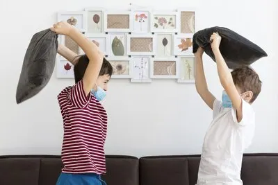 Side view of kids with medical masks playing with pillows at home