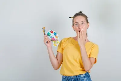 Young woman holding painting tools with hand on mouth in yellow t-shirt, jeans shorts and looking surprised