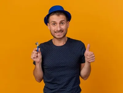 Impressed handsome man wearing blue party hat thumbs up and holds party whistle isolated on orange wall with copy space