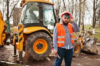 Brutal beard worker man suit construction worker in safety orange helmet sunglasses against traktor with adjustable wrench at hand