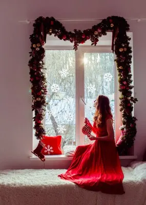 Lady in red dress sits on the widnowsill decorated with Christmas garland