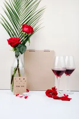 Side view of red roses with palm leaf in a glass bottle standing near a sketchbook and two glasses of red wine on white background