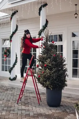 Pretty lady in red sweater standing on step- ladder while decorating Christmas tree in the yard