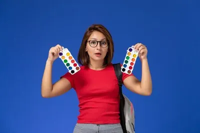 Front view of female student in red shirt with backpack holding paints for drawing on blue wall