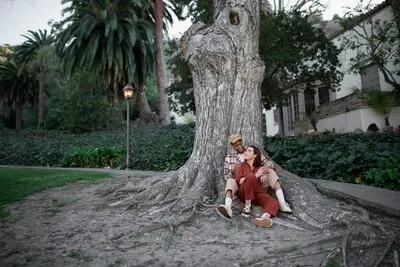 Trans couple holding hands and looking into each other's eyes while sitting by a big tree at the park