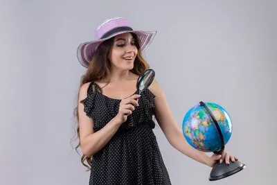 Young beautiful traveler girl in dress in polka dot in summer hat looking through magnifying glass with smile on face standing over white background
