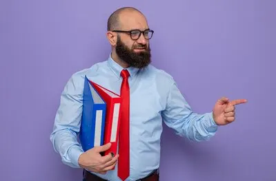 Bearded man in red tie and blue shirt wearing glasses holding office folders looking aside with smile on face pointing with index finger to the side