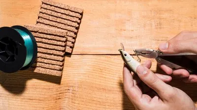 Close-up of a man fixing the hook with plier near the reel and cork board on wooden desk