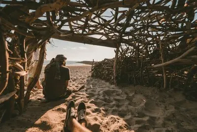 Person sitting inside a wooden cave taking a picture of a person sitting on the sand near the sea