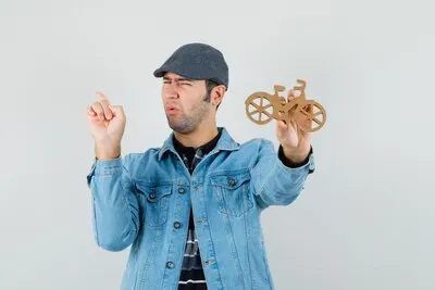 Young man pointing back, holding wooden toy bike in t-shirt, jacket, cap and looking discontent. front view.