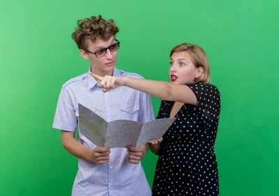 Young beautiful couple man holding map looking at his confused girlfriend which pointing at something with finger standing over green wall