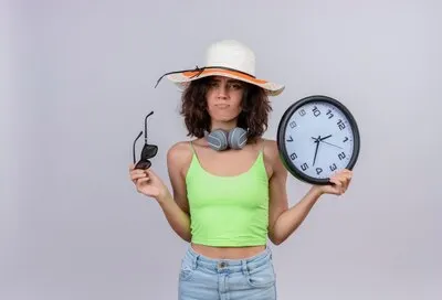 A displeased young woman with short hair in green crop top wearing sun hat holding sunglasses and wall clock on a white background