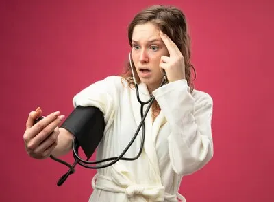 Scared young ill girl wearing white robe measuring her own pressure with sphygmomanometer putting finger on temple isolated on pink
