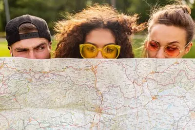 Happy young funny company of friends tourists hiding behind map in sunglasses, man and women having fun together, traveling
