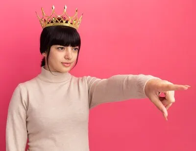Confident young brunette caucasian girl with crown looks and points at side on pink