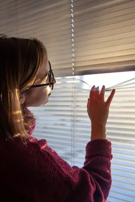 A woman looks through the blinds at the early morning sunlight