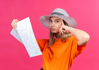 A confused pretty young woman in an orange t-shirt wearing sunhat holding map while pointing at head and looking side on a pink wall