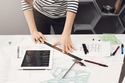 Top view of young good-looking architect student girl in casual striped shirt and black jeans standing near table, holding ruler and pen in hands making drawings, watching movie on digital table, gett
