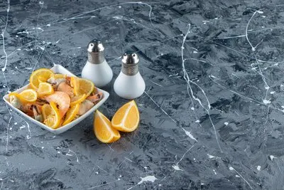 Sliced lemons and prawns in a bowl next to salt, on the marble background.