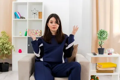 Clueless young pretty caucasian girl sitting on armchair in designed living room  keeping hands in air