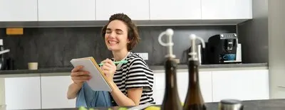 Close up of happy smiling young woman writing in notebook creating recipe list of meals for this