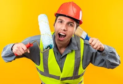 Excited young builder man in uniform holding roller brush with paint brush isolated on yellow wall