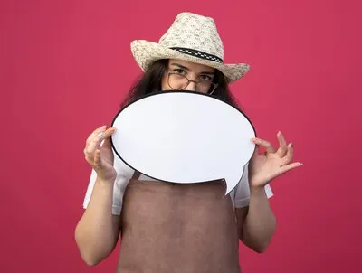 Confident young brunette female gardener in optical glasses and in uniform wearing gardening hat holds speech bubble isolated on pink wall