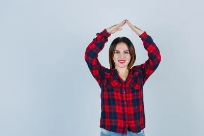 Young woman making house roof gesture over head in checked shirt and looking cheerful. front view.