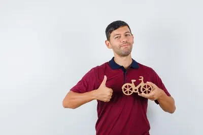 Young man in t-shirt holding wooden toy bike with thumb up and looking satisfied , front view.